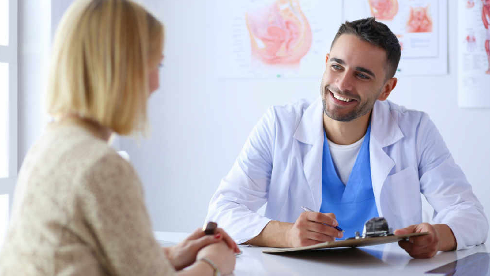 A young male doctor, holding a clipboard and smiling, while sat at a table with a blonde female patient